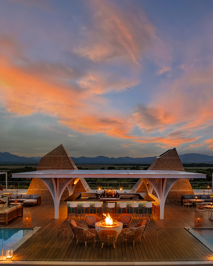 A stylish outdoor lounge with wicker chairs around a fire pit, a bar under a modern white pergola, and unique pyramid-shaped structures, set against a colorful sunset sky and mountain backdrop.