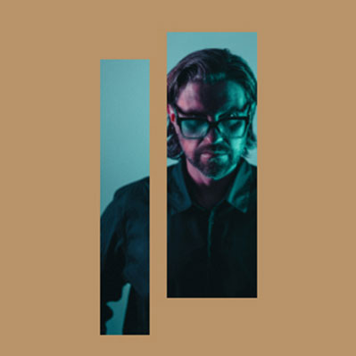 Un hombre con el pelo hasta los hombros y gafas está iluminado con luz verde azulado y violeta. Está parcialmente oculto por dos barras verticales de color canela sobre un fondo canela.