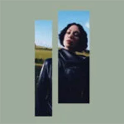 Una mujer con el pelo corto y rizado está de pie al aire libre contra un cielo azul y un campo verde, enmarcada por dos recortes rectangulares verticales sobre un fondo verde apagado.