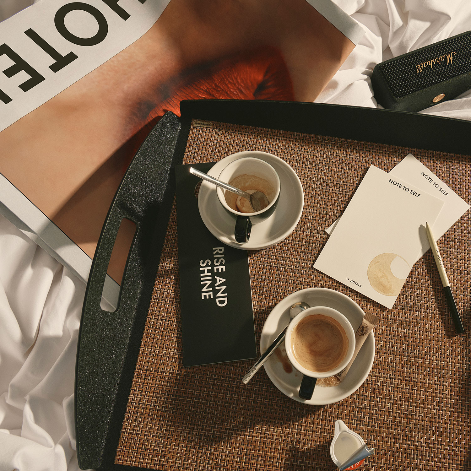 A tray on a bed holds two cups of coffee, a spoon, a pen, papers, and a booklet titled “Rise and Shine.” Nearby are a magazine with lips on the cover and a black wallet. Soft natural light streams in.