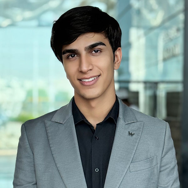 A young man with dark hair wearing a grey blazer over a black shirt, smiling while standing indoors in a modern, bright setting.