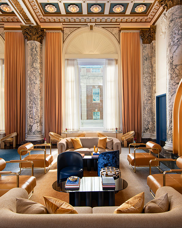 Elegant lounge with high, ornate ceiling, marble columns, tall arched windows with gold curtains, plush beige sofas, gold armchairs, and modern coffee tables under natural light.