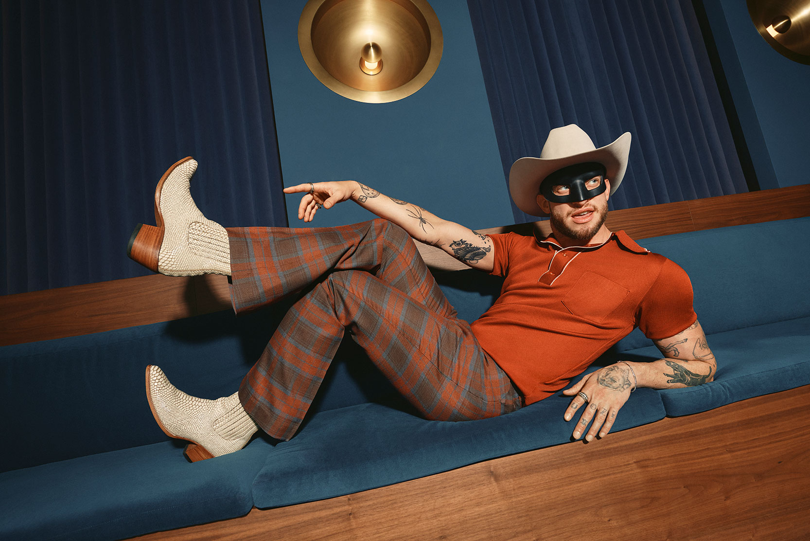 A man wearing a white cowboy hat, a black eye mask, an orange shirt, plaid flared pants, and snakeskin boots lounges on a blue sofa, pointing playfully. The background features blue walls and gold circular lights.