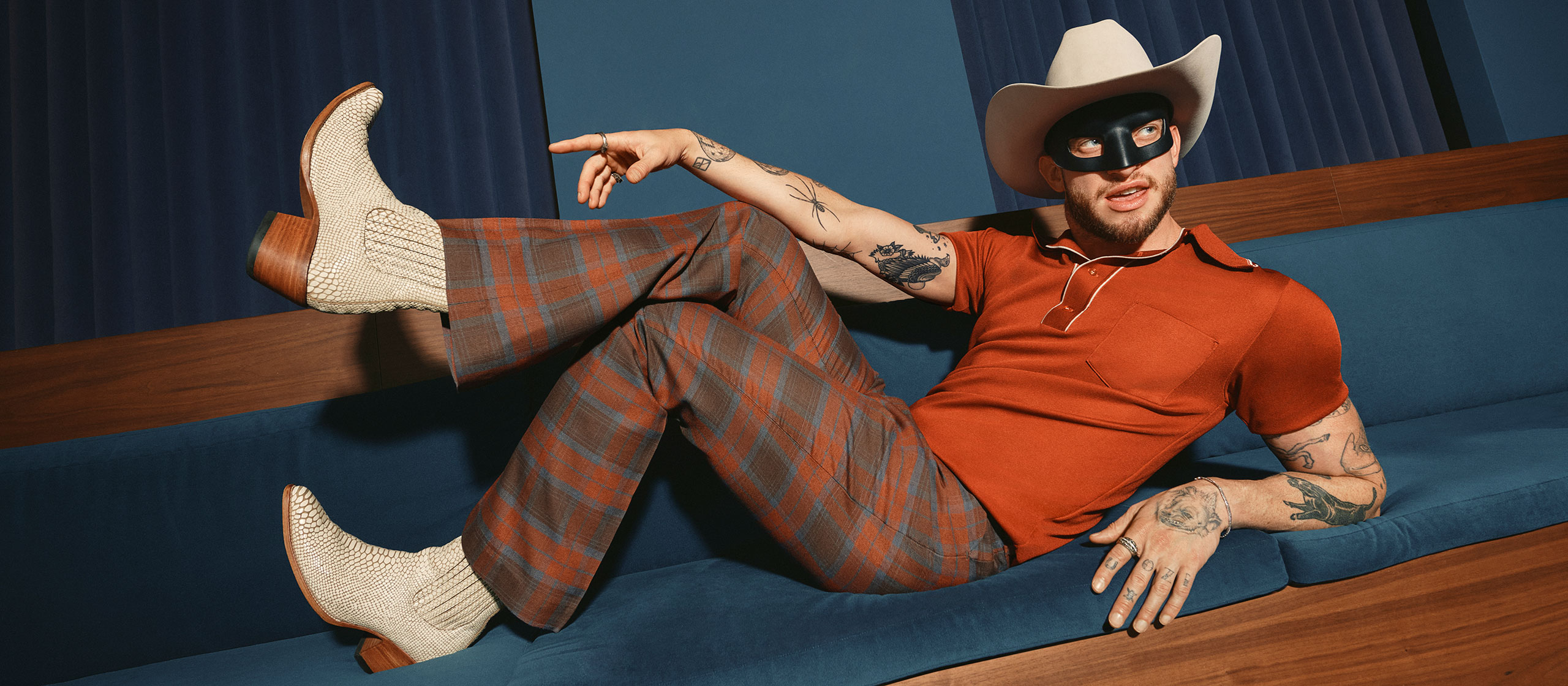 A man wearing a cowboy hat, black eye mask, orange polo shirt, plaid pants, and white boots lounges on a blue sofa, striking a playful pose with one leg raised and pointing to the side.