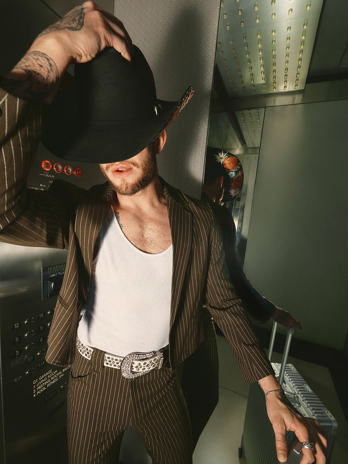 A person in a pinstripe suit and white tank top stands in an elevator, tilting a black cowboy hat to partially cover their face. They wear a decorative belt and have tattoos on their arm. Another person is reflected in the mirror.