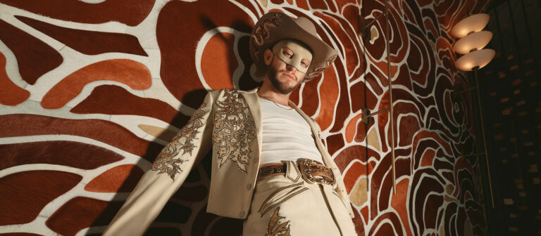 A man in a decorated cream suit, cowboy hat, and mask poses against a bold, abstract brown and white patterned wall, looking confidently down at the camera.