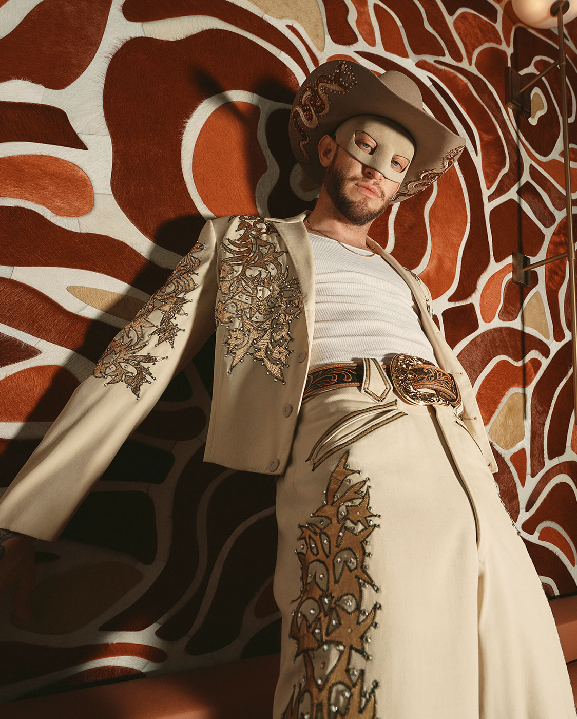 A person in a decorated cream suit, white tank top, and cowboy hat leans against a retro-patterned wall. They wear a tan mask and ornate belt, creating a bold, stylish look in a vintage-inspired setting.