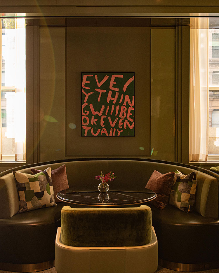A cozy curved booth with patterned pillows surrounds a round black table with a small flower vase. Behind it, abstract art on the wall reads “Everything will be ok eventually.” Sunlight filters through tall windows.