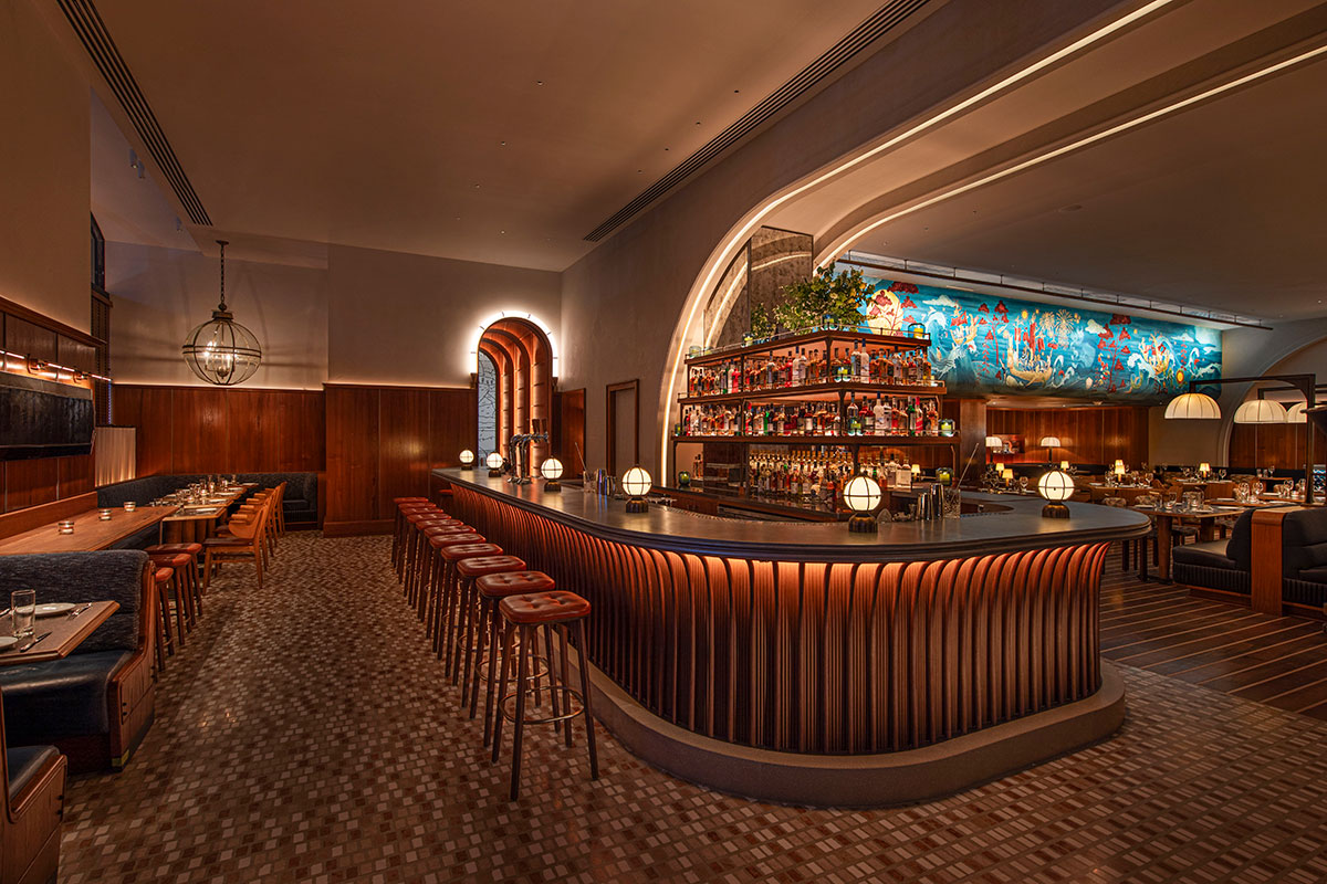 A stylish, dimly lit restaurant bar with curved wooden counters, leather bar stools, art deco lighting, and a colorful mural above the back bar. There are booth and table seating areas along the walls.