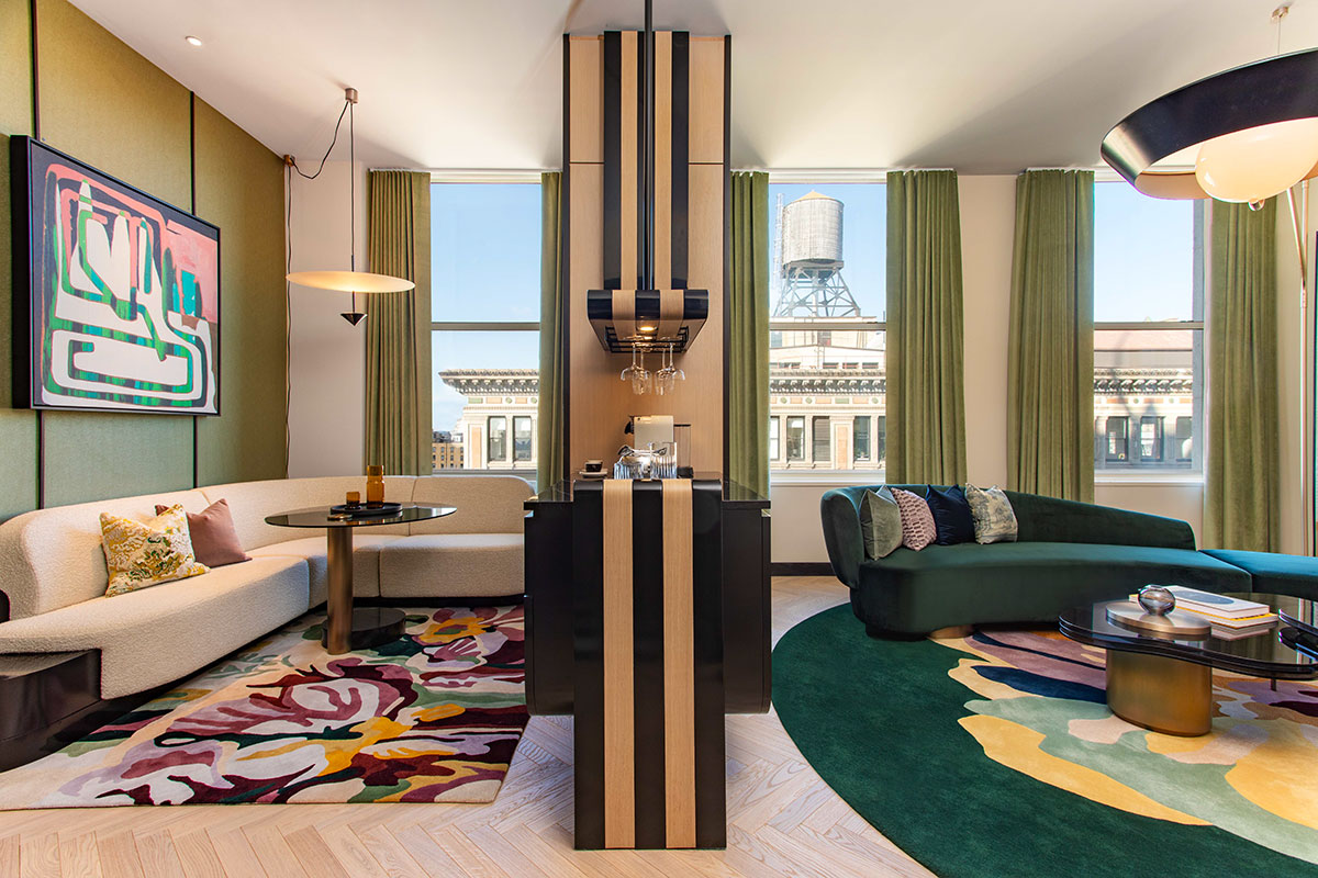 A stylish living room with modern furniture, colorful rugs, green curtains, large windows, and a view of city buildings and a water tower outside. The room features a partition with striped wood and a built-in bar area.
