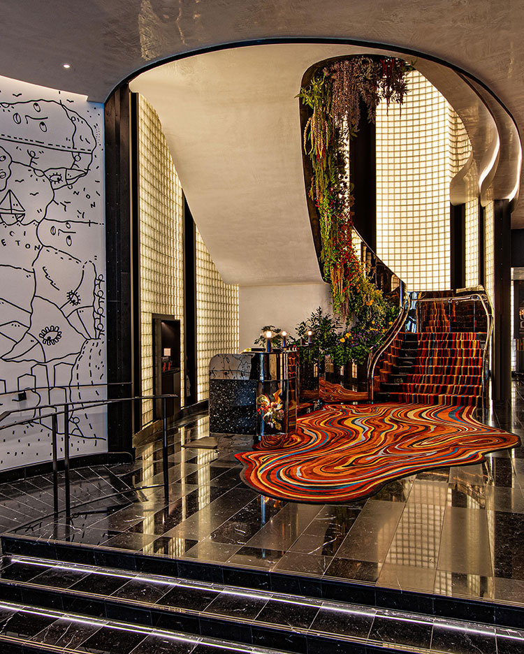 Modern hotel lobby with black marble floors, a colorful wavy rug, a curved staircase adorned with greenery, glass block walls, and a white mural with abstract line art on the left wall.