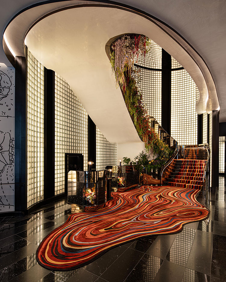 A grand staircase with lush greenery and flowers along the railing curves upward, surrounded by glass brick walls. Below, a vibrant red, orange, and purple patterned carpet flows onto glossy black floors.
