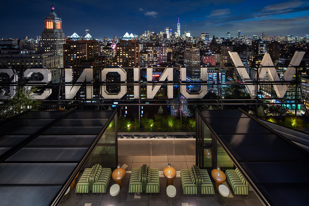 Rooftop bar with striped chairs and round tables overlooks a vibrant city skyline at night; large illuminated “W UNION SQ” sign is visible, with skyscrapers and city lights in the background.