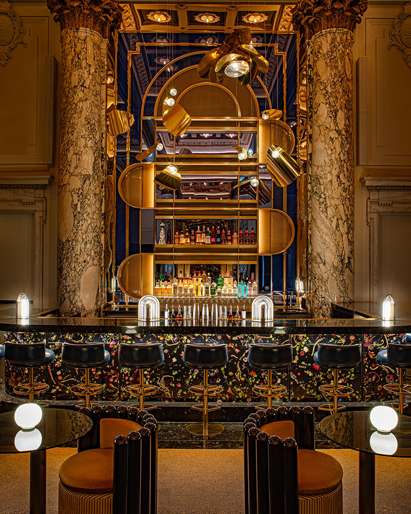 A luxurious bar with marble columns, ornate ceiling, and modern gold light fixtures. The bar counter has a floral pattern and black stools, with shelves of colorful bottles and elegant lighting throughout.