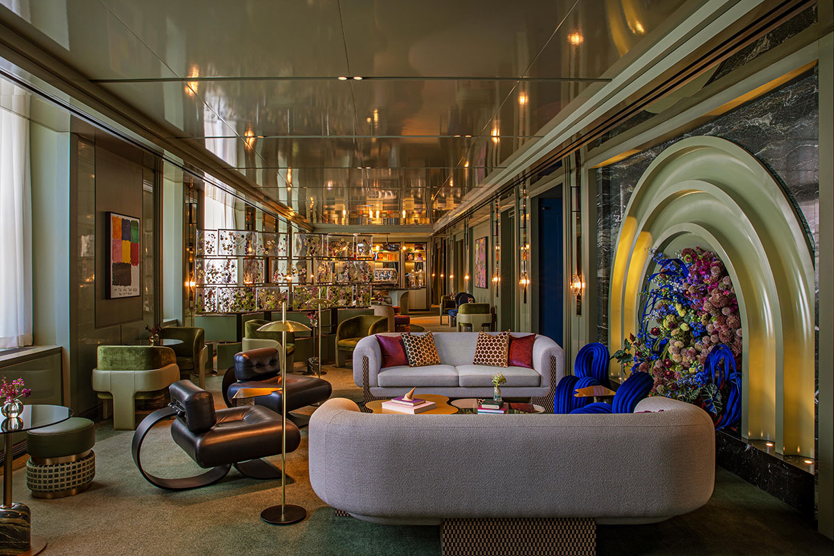 A luxurious, modern lounge with plush sofas, unique black chairs, colorful cushions, floral art, and stylish lighting. The space features a well-lit bar in the background and a dramatic flower arrangement on the right wall.