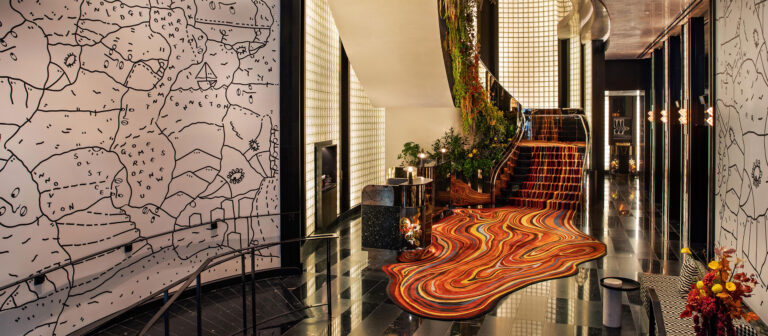 A stylish hotel lobby features glossy black floors, a wavy red-orange carpet, a plant-adorned staircase, and walls with abstract map-like drawings. Modern lighting and floral arrangements add to the vibrant atmosphere.
