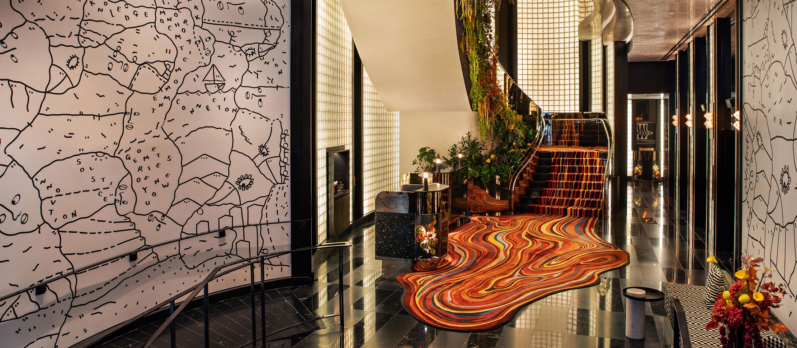 A stylish hotel lobby features glossy black floors, a wavy red-orange carpet, a plant-adorned staircase, and walls with abstract map-like drawings. Modern lighting and floral arrangements add to the vibrant atmosphere.