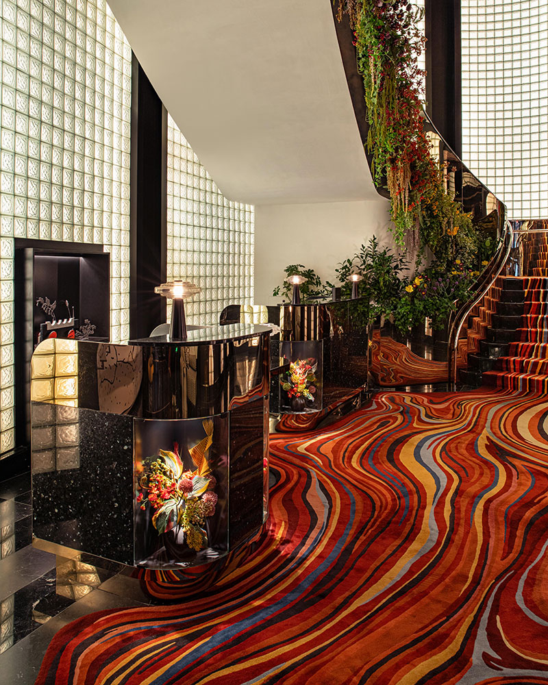 Reception area with glass block walls, glossy black reception desks, vibrant red and orange wavy carpet, and a staircase decorated with cascading plants and flowers. Warm lighting creates a modern, luxurious atmosphere.