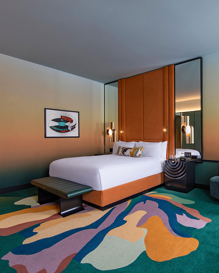 Modern hotel room with a tall orange headboard, white bed, colorful abstract carpet, wall art, two bedside tables, brass wall sconces, and mirrors behind the bed, creating a stylish and vibrant atmosphere.