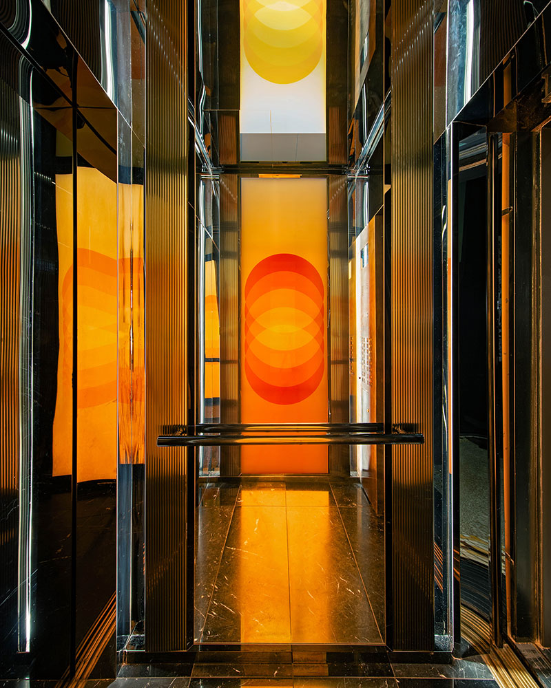 A modern elevator interior with glossy black walls and floor, featuring bright orange and yellow overlapping circles as illuminated wall art on the back panel, creating a warm, vibrant atmosphere.