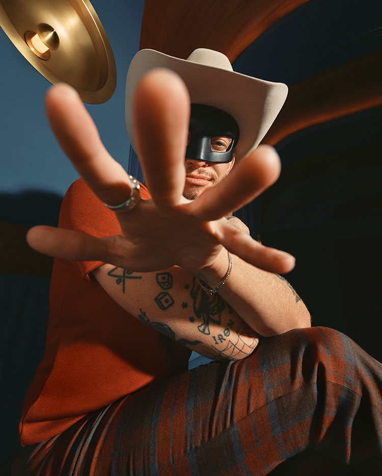 A person wearing a white cowboy hat, a black mask, and red shirt reaches toward the camera, their hand outstretched. Tattoos are visible on their arm, and they are dressed in plaid pants. The background is dramatic and dimly lit.