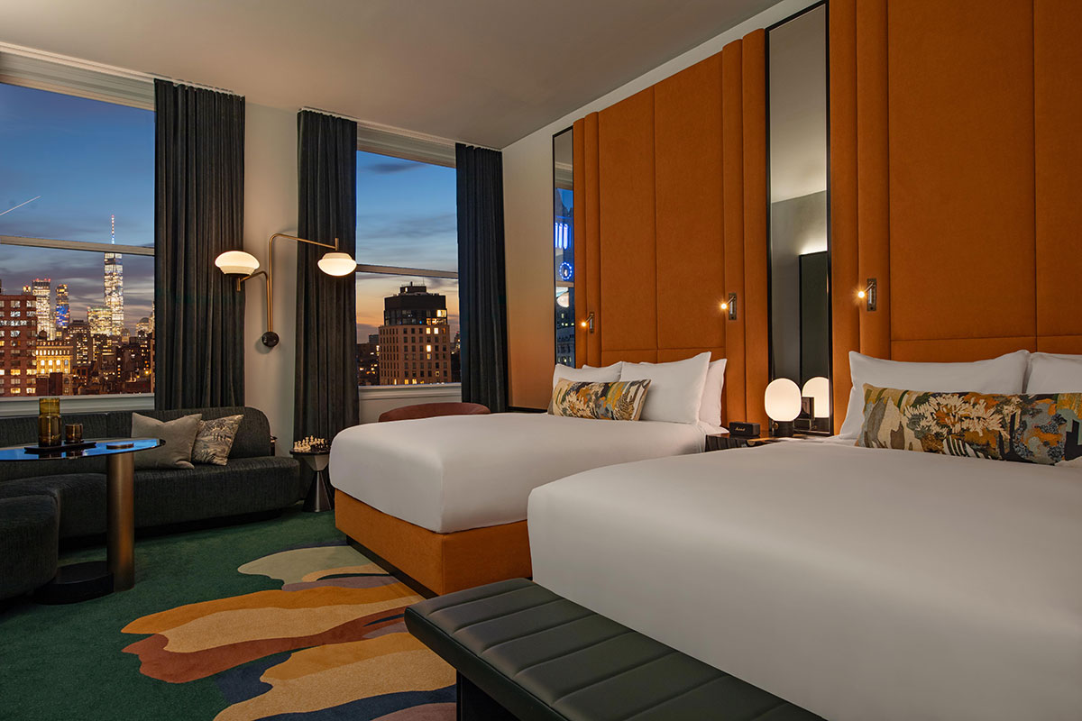 Modern hotel room with two large beds, stylish orange and brown decor, patterned rug, dark sofa, round table, floor lamp, and large windows showing a city skyline at dusk.