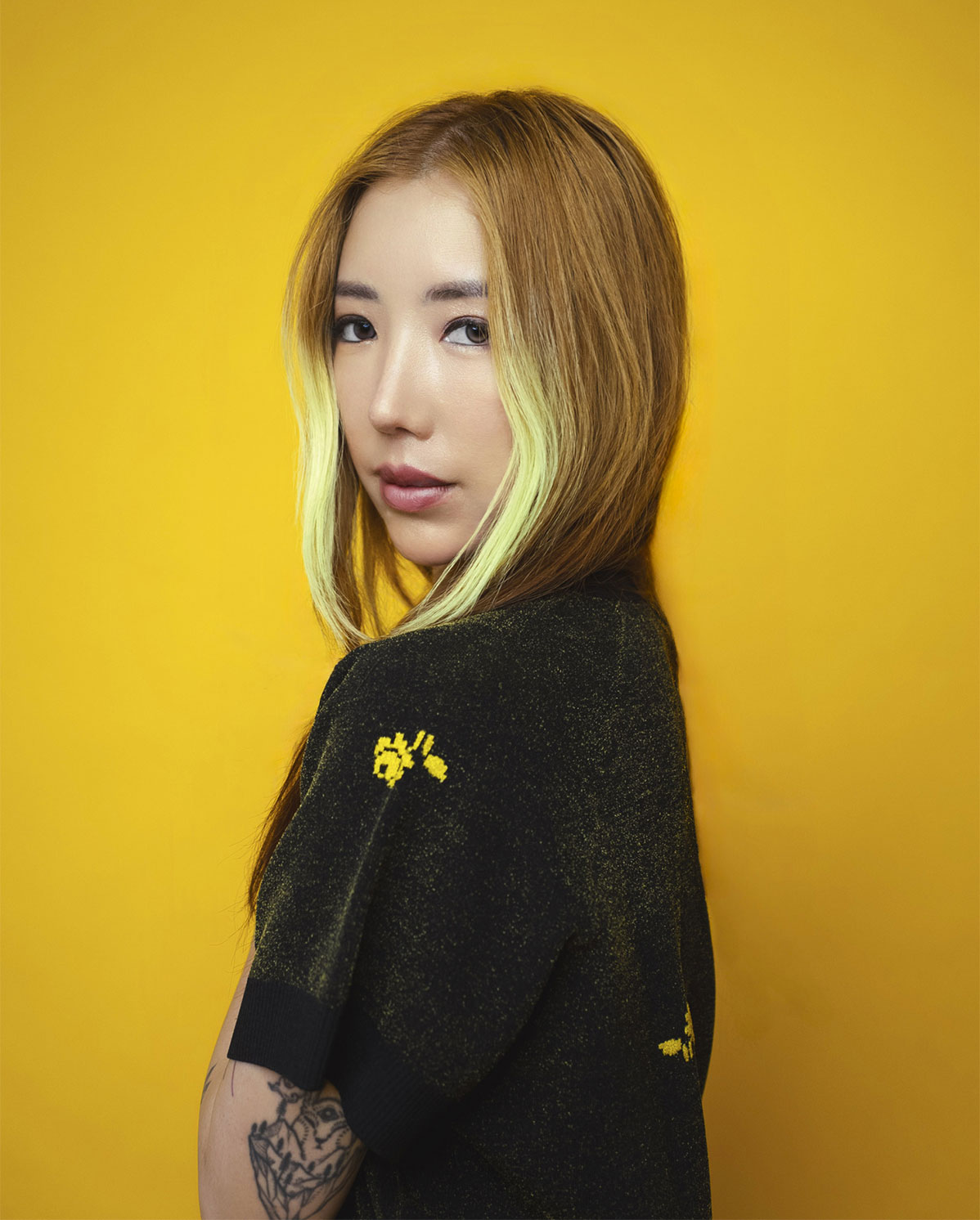 A person with long, light brown hair featuring green highlights poses against a vibrant yellow background, wearing a black shirt with yellow speckles—W PRESENTS this striking look while they reveal a bold tattoo on their arm.