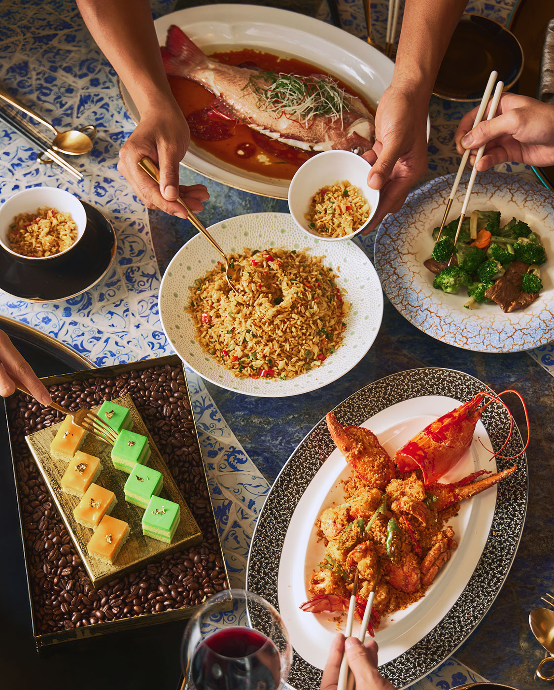 A table set with various Chinese dishes, including whole fish, fried rice, lobster, beef with broccoli, a tray of green and yellow desserts, and bowls of crispy topping, with several hands serving and eating.
