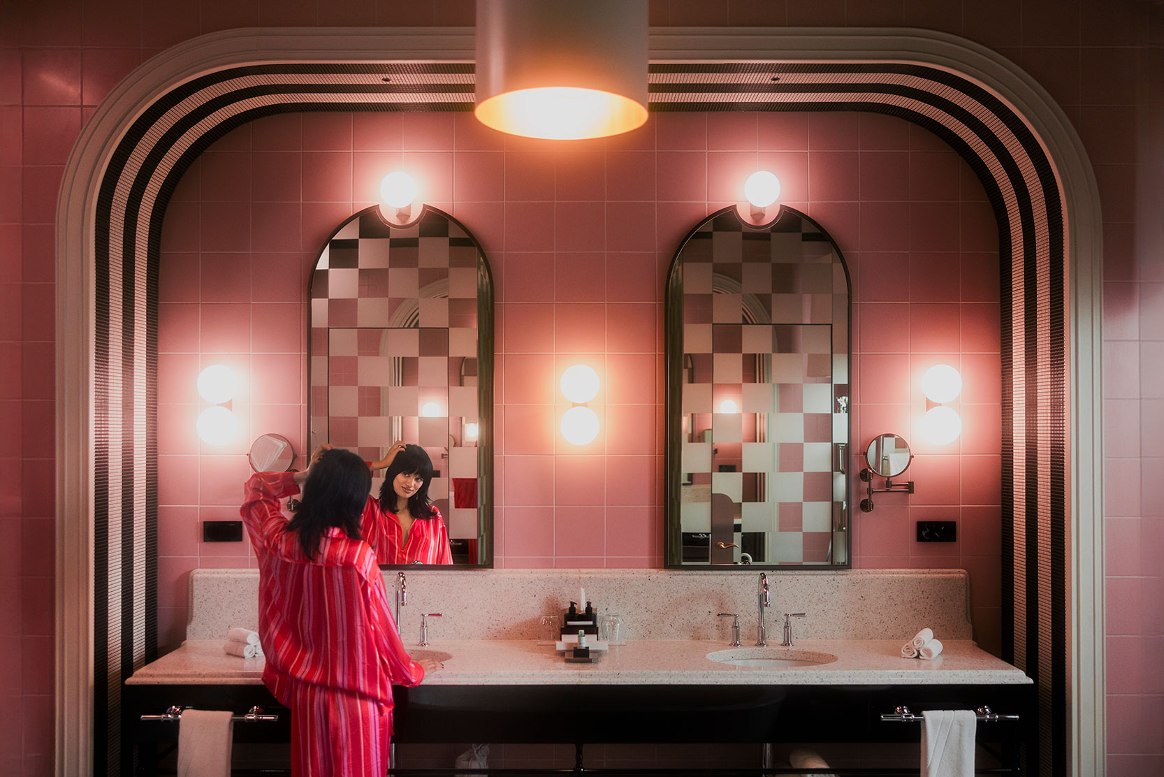 A person in a pink striped robe stands at a bathroom sink with pink tiled walls, looking at their reflection in a large arched mirror. The room is warmly lit with round wall lights above and between the mirrors.