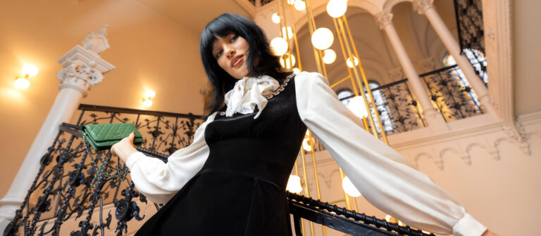 A woman in a black dress with a white blouse stands on a staircase with ornate black railings, holding a green clutch. Warm lights and decorative columns are visible in the background.