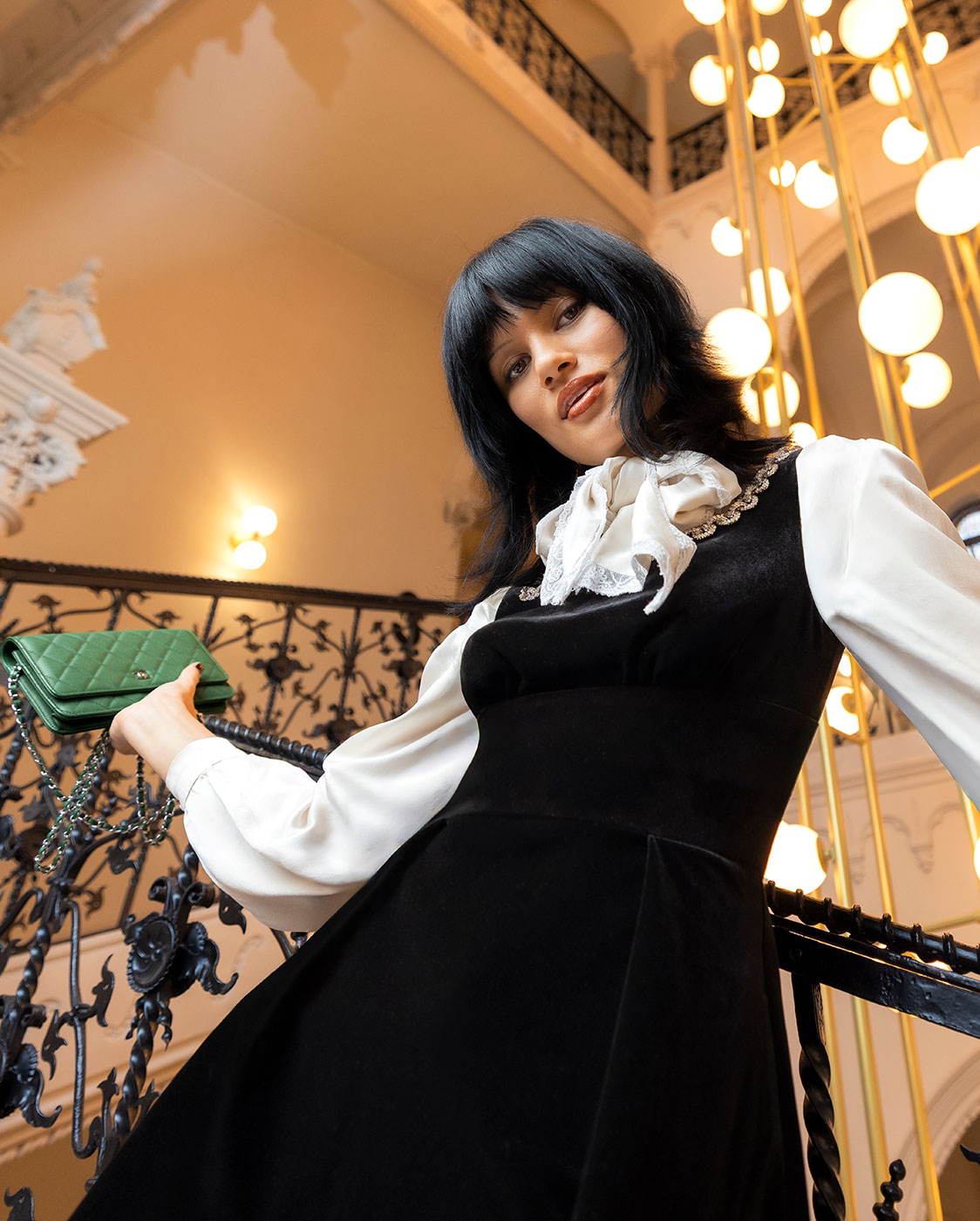 A woman with black hair, wearing a white blouse with a bow and a black dress, poses on a decorative staircase holding a green quilted handbag. Spherical lights and elegant railings are visible in the background.