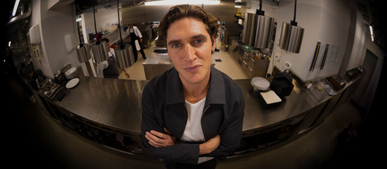A man with wavy brown hair stands confidently with arms crossed in a modern, stainless steel commercial kitchen, looking directly at the camera. Several people work in the background.