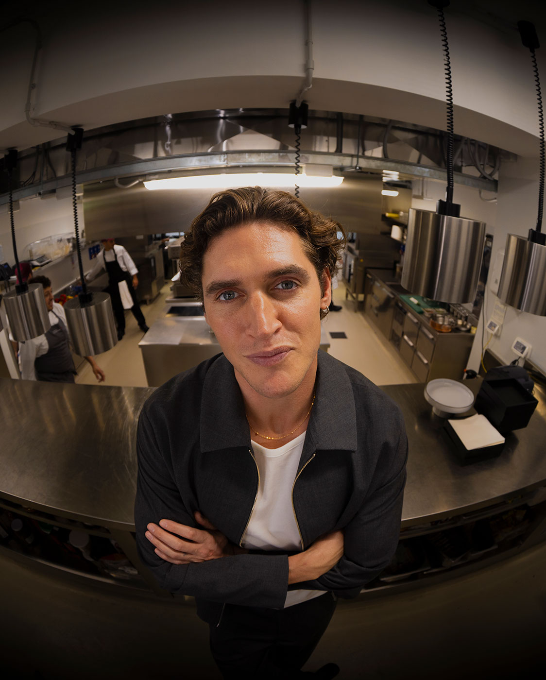 A man with curly brown hair and blue eyes stands in a commercial kitchen, looking directly at the camera with his arms crossed. Stainless steel appliances and countertops are visible in the background.
