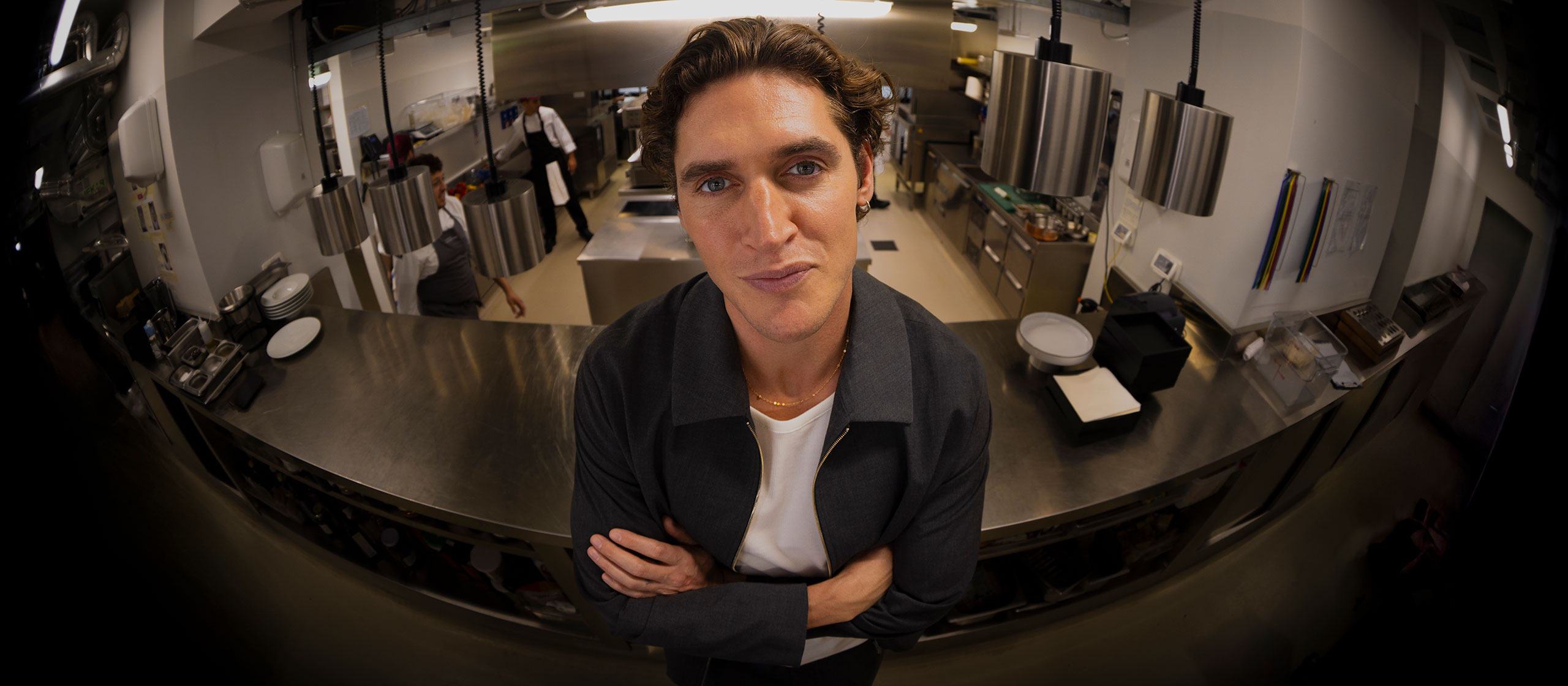 A man with wavy brown hair stands confidently with arms crossed in a modern, stainless steel commercial kitchen, looking directly at the camera. Several people work in the background.