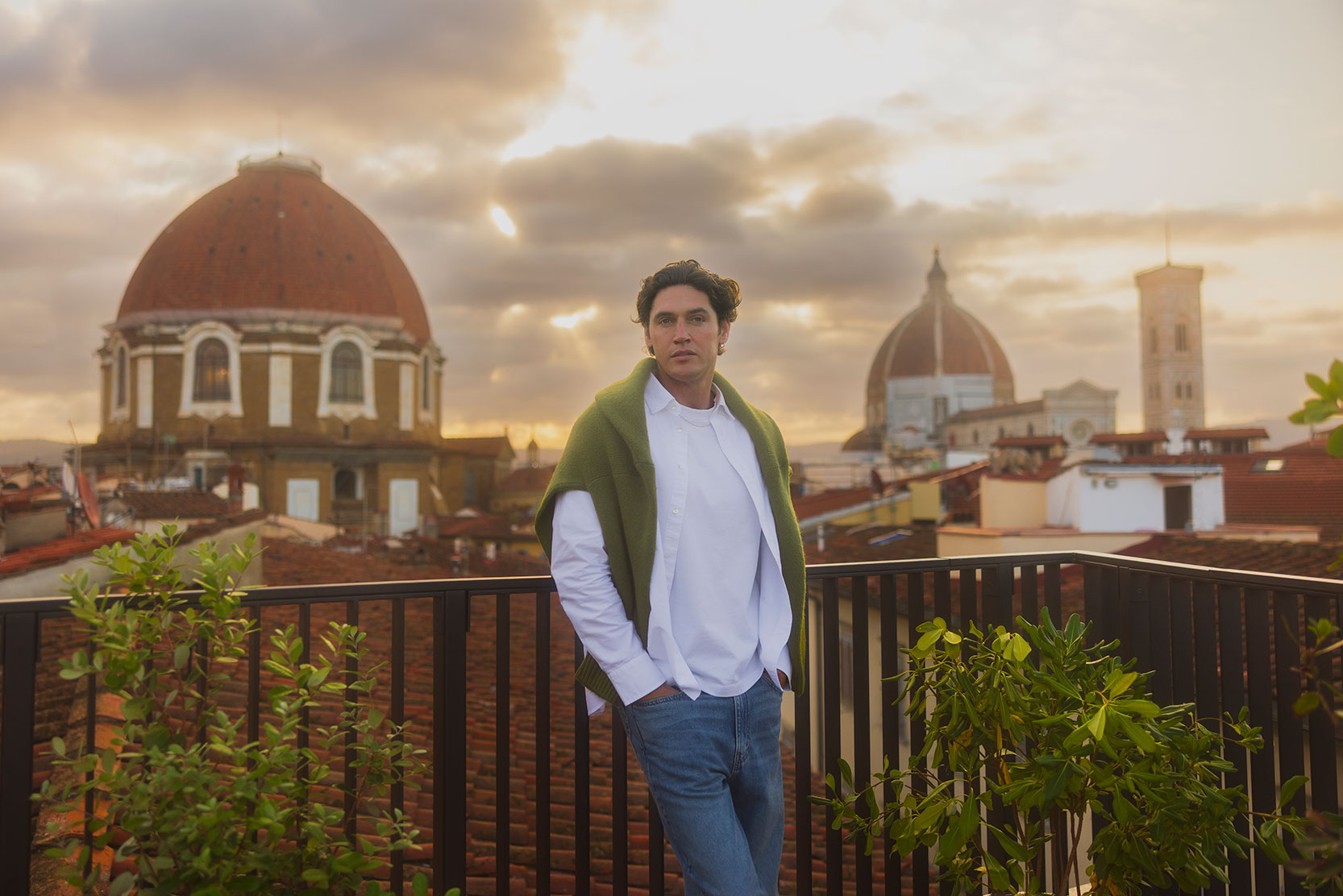 A person in a white shirt and green sweater stands on a balcony with hands in pockets, overlooking Florence’s rooftops with the iconic domes of the city and a cloudy sunset sky in the background.