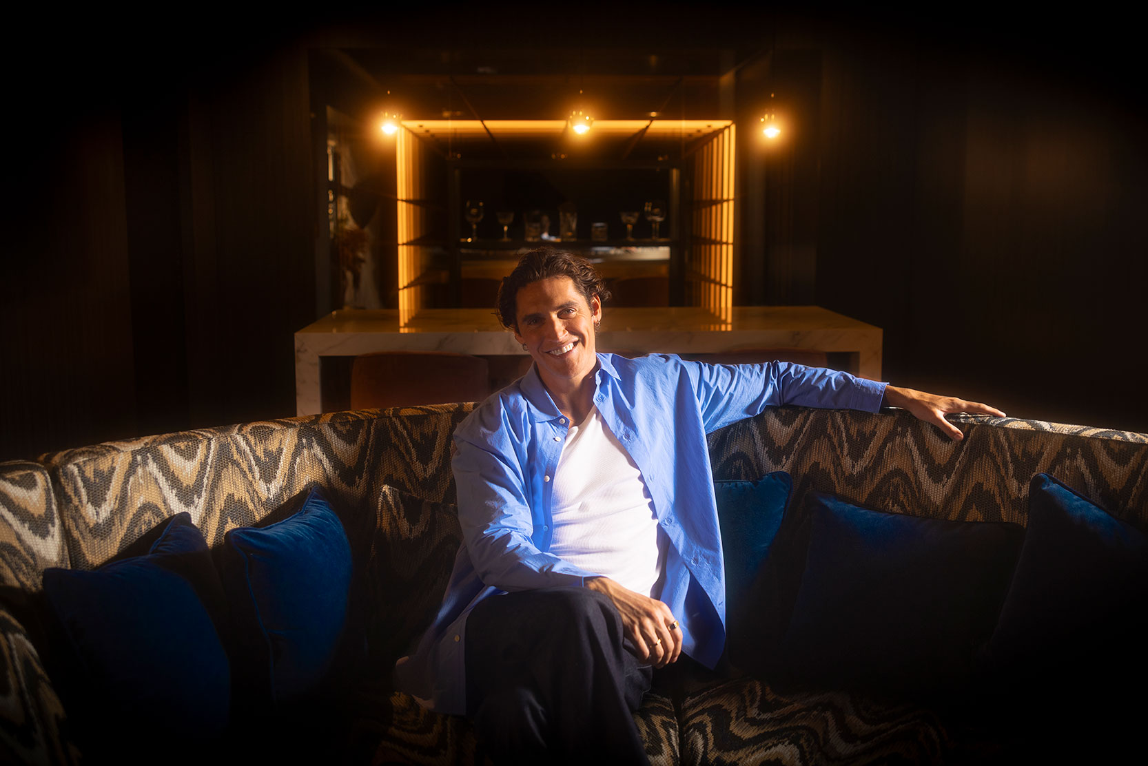 A man in a blue shirt and white t-shirt sits smiling on a patterned sofa with blue cushions in a dimly lit, stylish lounge with a glowing bar in the background.