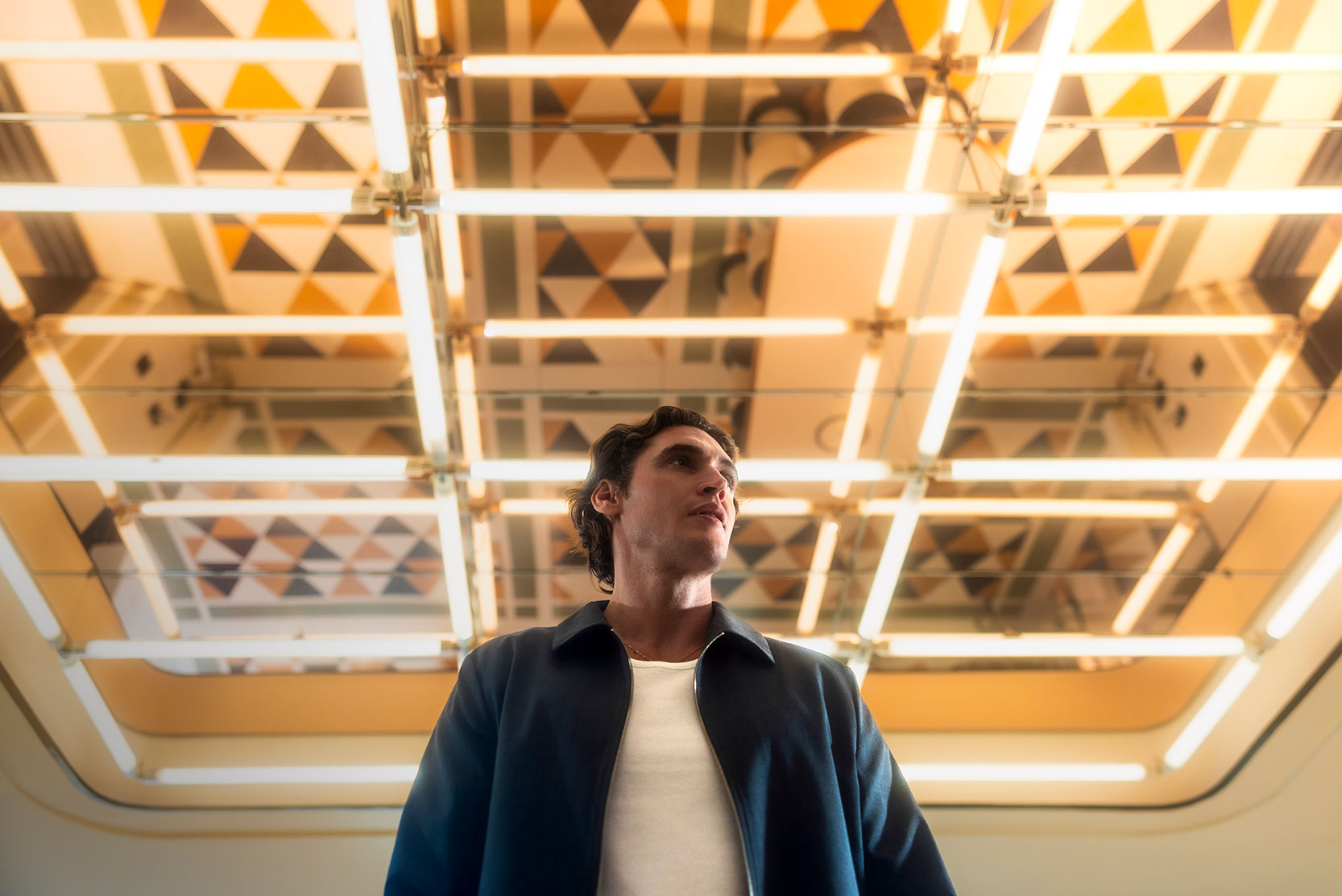 A person in a dark jacket and white shirt stands under a ceiling with geometric patterns and bright fluorescent lights, looking slightly to the side. The scene has a modern, futuristic atmosphere.
