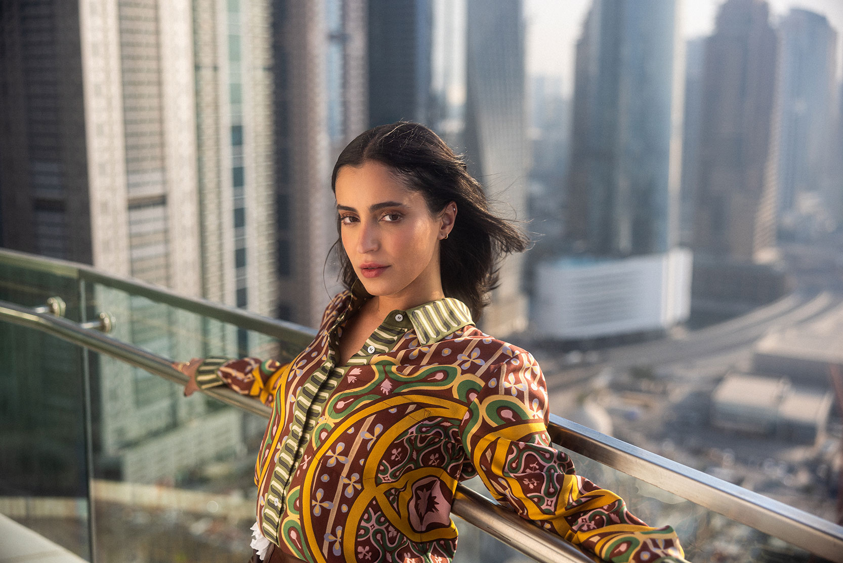 A woman with long dark hair leans against a glass balcony railing, wearing a colorful patterned jacket. Tall skyscrapers and a cityscape are visible in the background on a bright, hazy day.