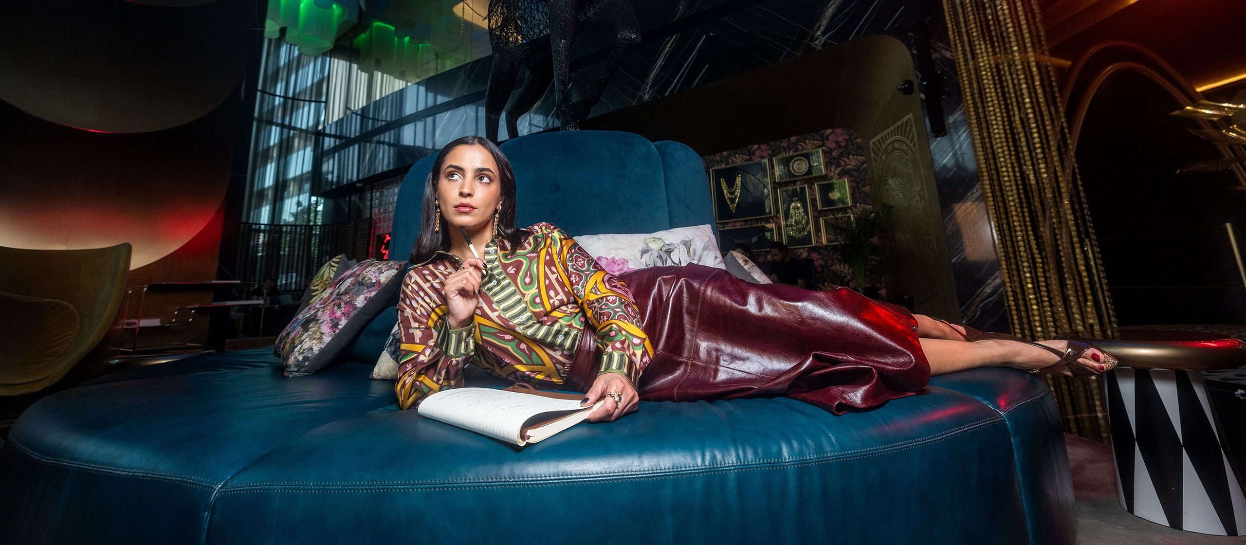 A woman in a colorful blouse and maroon skirt lounges on a large, round blue sofa, holding a pen and an open notebook, in a stylish, art-filled modern room.