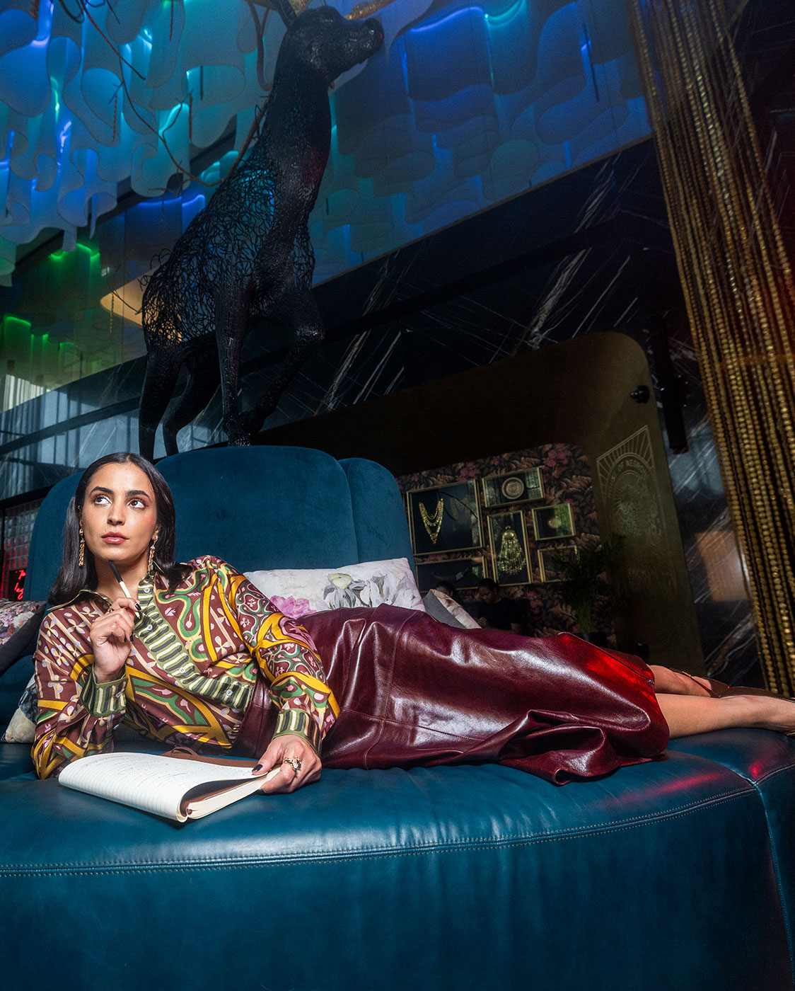 A woman in a colorful blouse and long maroon skirt lounges on a large blue chair, holding a pen and an open notebook, with a statue of a deer behind her in a modern, stylish room.