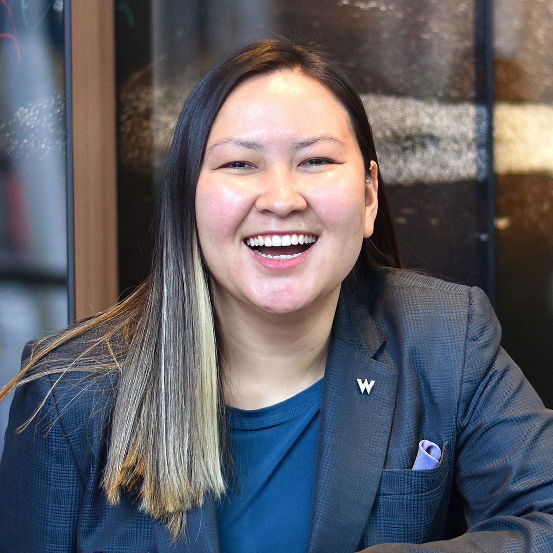 Eine Person mit langem, glattem Haar und einer blonden Strähne lächelt, trägt ein blaues Hemd und einen grauen Blazer mit einer 