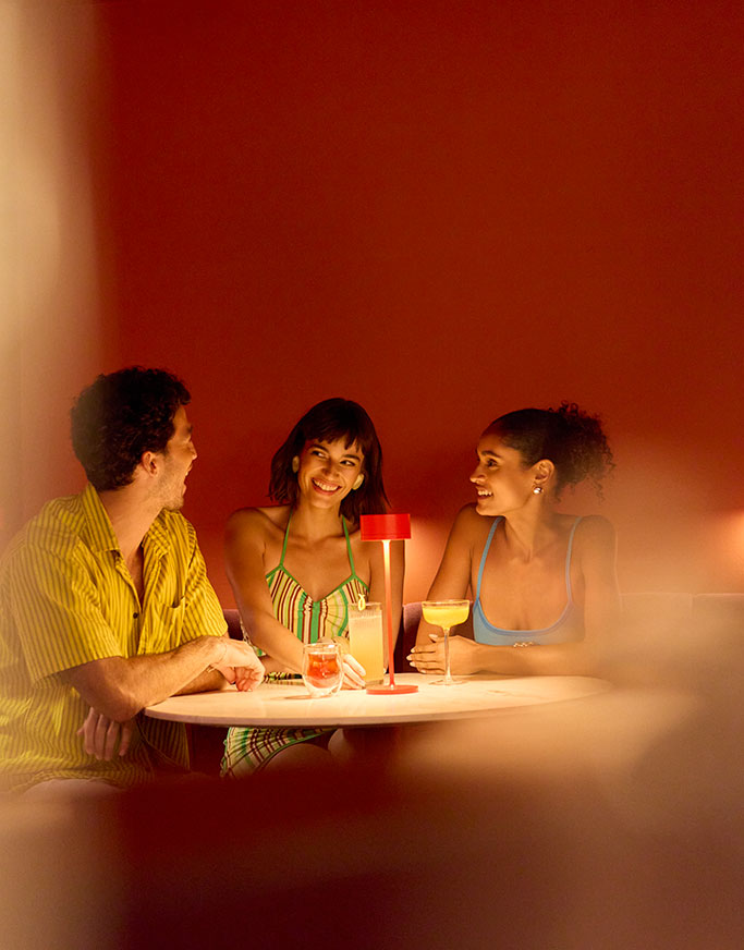Three people sit around a small round table in a warmly lit room, smiling and talking. Drinks are on the table and a red lamp sits in the center, illuminating their faces. The background is a solid deep orange color.