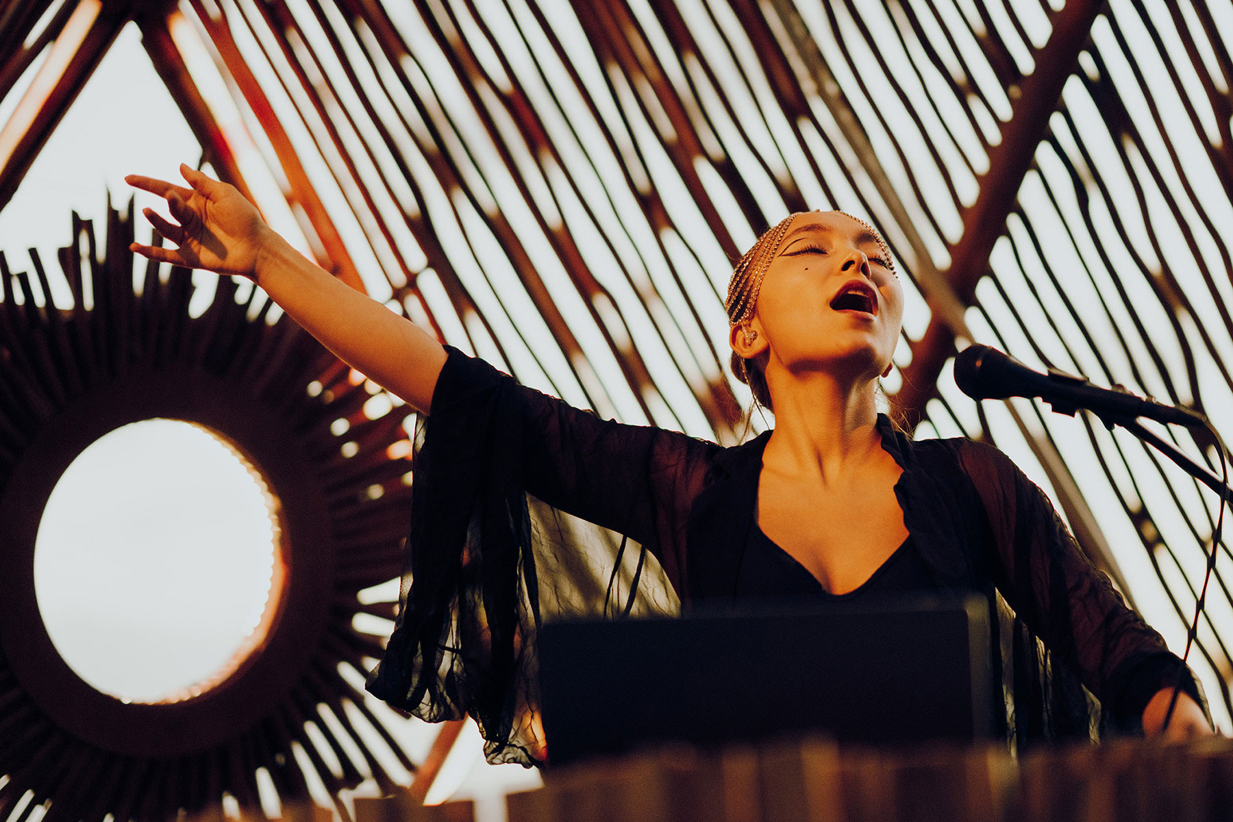 A woman passionately sings into a microphone with one arm raised, standing in front of a decorative, sun-shaped wooden backdrop with radiating beams. Warm light highlights her expressive face and dramatic pose.