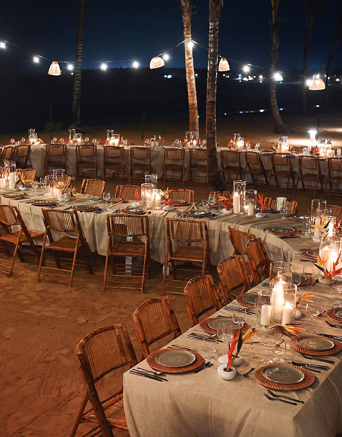 An outdoor dining area set up on sand at night, with long tables covered in beige cloth, wooden chairs, candles, plates, glasses, and string lights creating a warm, festive atmosphere.