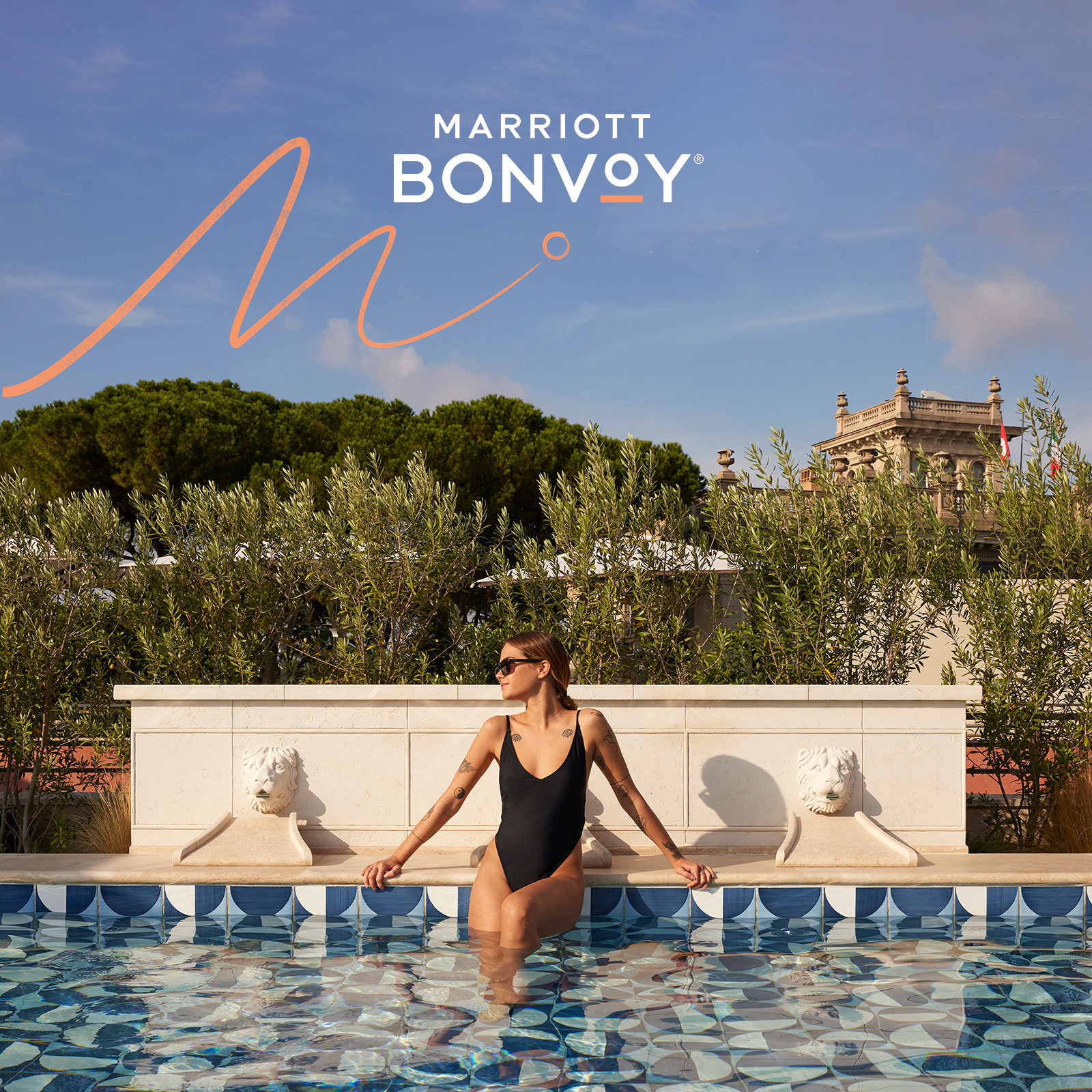 A woman in a black swimsuit relaxes on the edge of a decorative outdoor pool with a patterned tile design. Olive trees and classical architecture are in the background. 