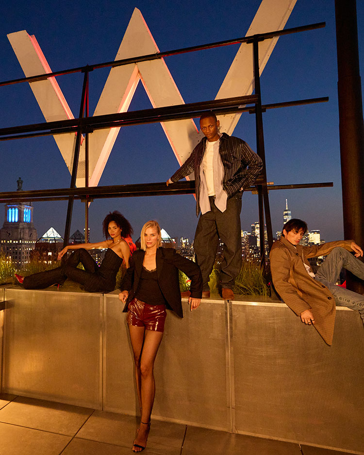 Four stylishly dressed people pose confidently at night on a rooftop in front of a large illuminated 