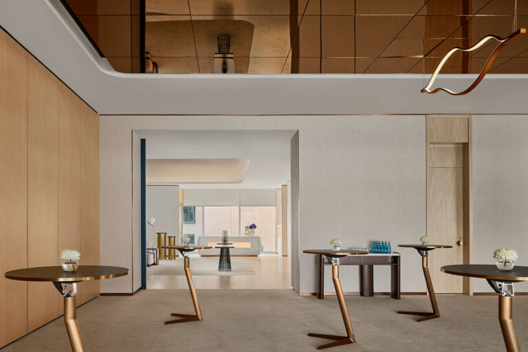 A modern interior with round cocktail tables, light wood walls, neutral carpeting, minimalist wall art, and a reflective ceiling. Small vases with white flowers sit on the tables, and a stylish chandelier hangs above.