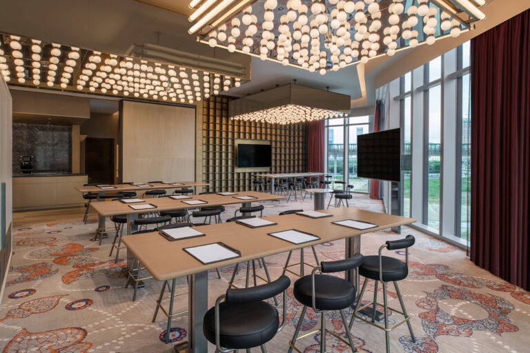 A modern conference room with large windows, geometric light fixtures, several tables with notepads and pens, black chairs, patterned carpet, and floor-to-ceiling red curtains.