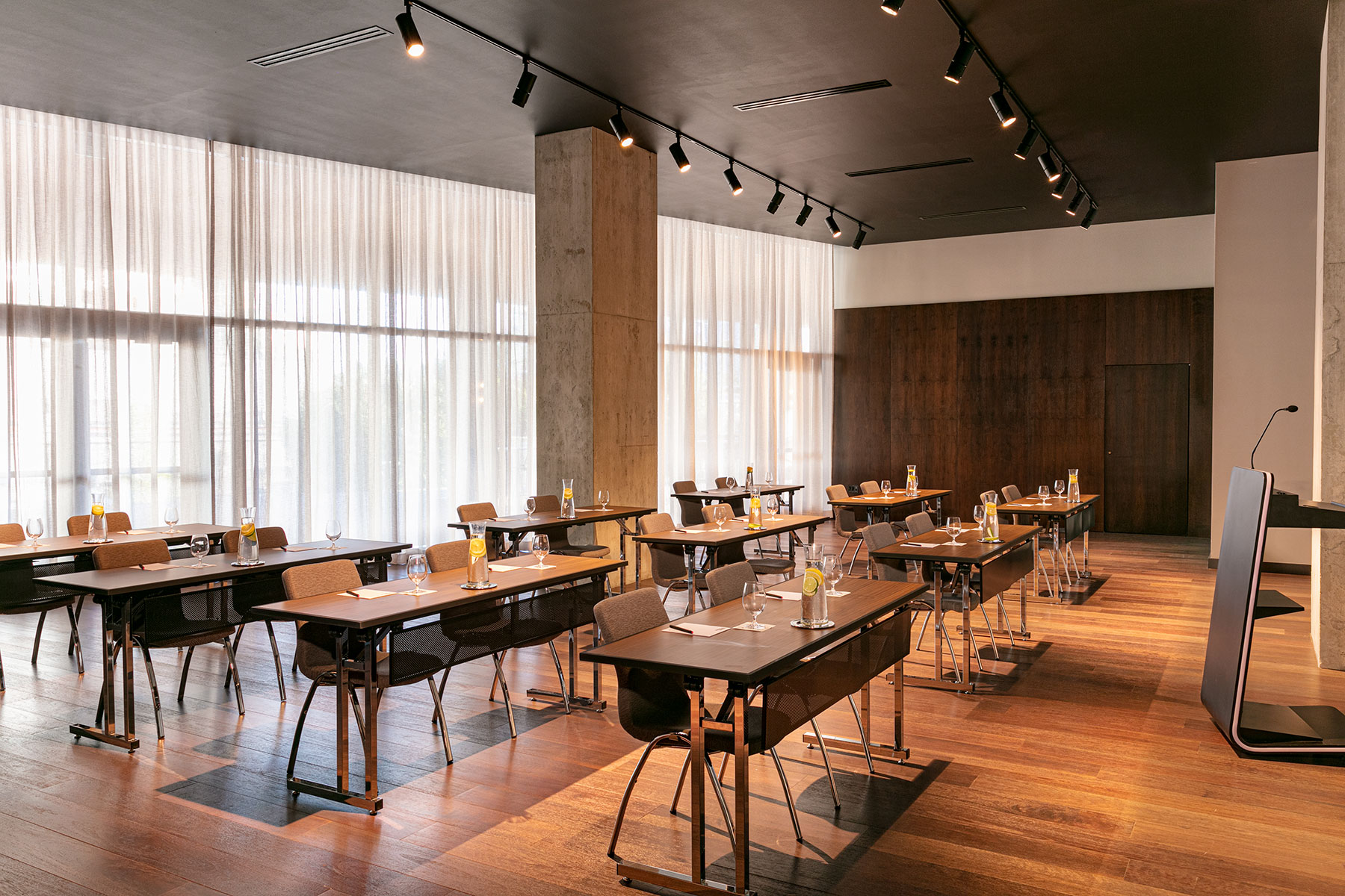 A modern conference room with several rows of tables and chairs, set up with water glasses, notepads, and pens. Large windows with sheer curtains let in natural light, and a podium stands at the front.