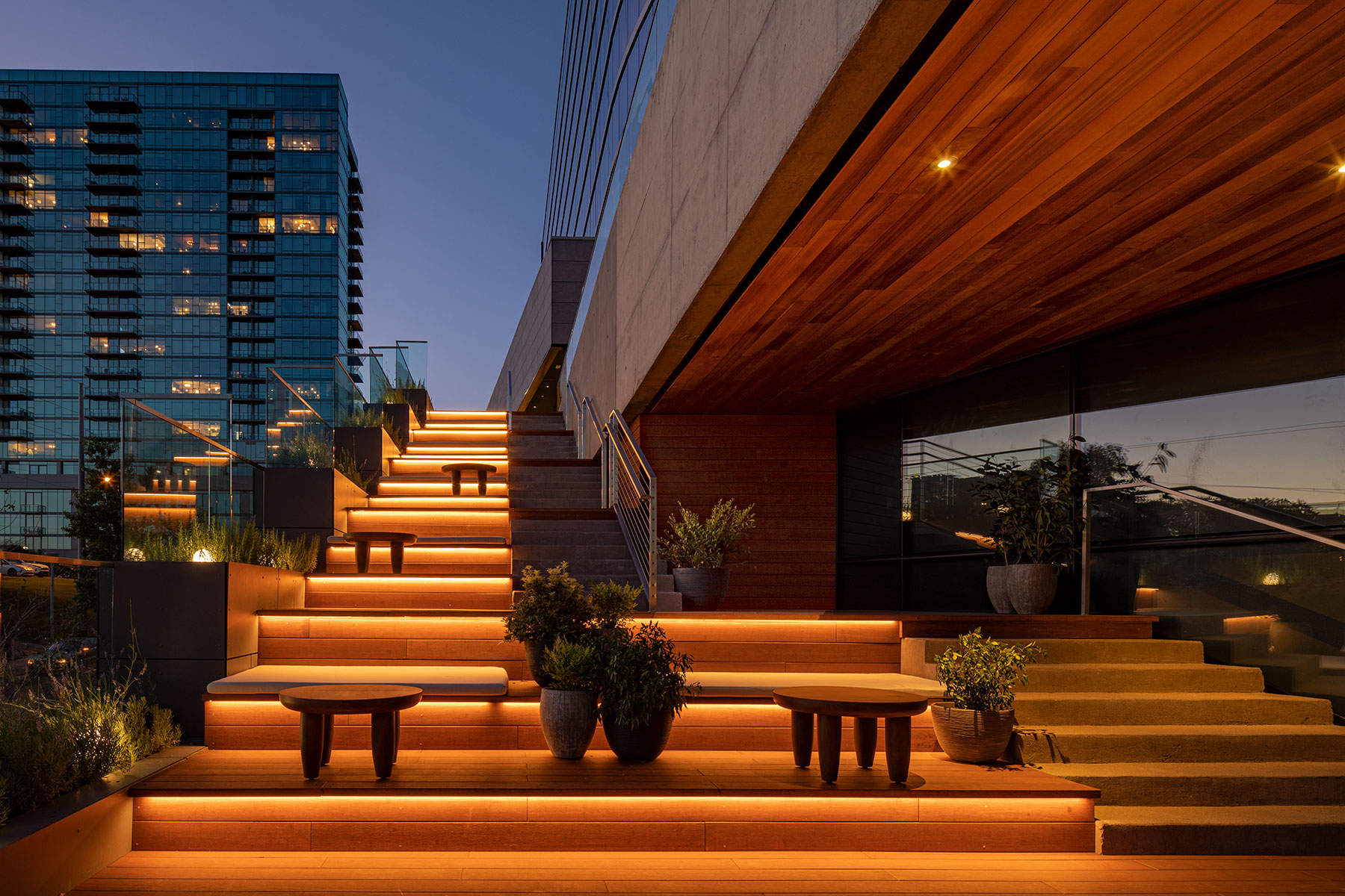 A modern outdoor staircase illuminated with warm, recessed lighting, surrounded by potted plants and seating areas, with a glass building in the background at dusk.